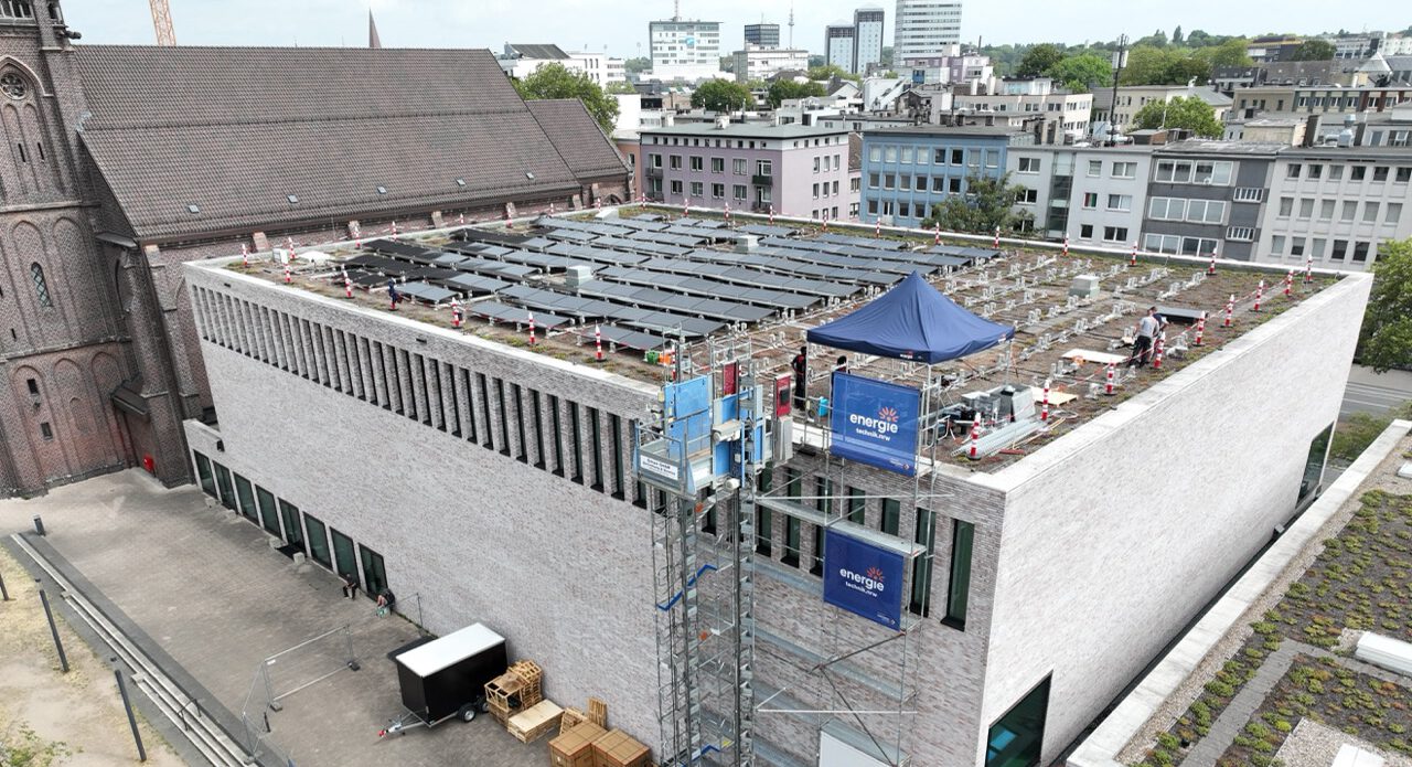 Aerial view of a flat-roofed building with rows of solar panels being installed; scaffolding, a blue canopy, and construction gear on the site.